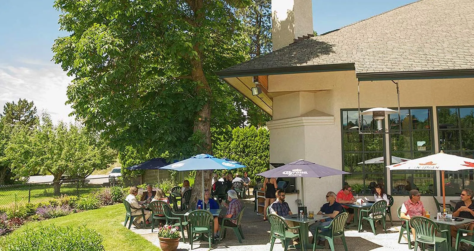 Wide view of McCulloch Station Pub's patio with guests relaxing under umbrellas and trees in summer sun
